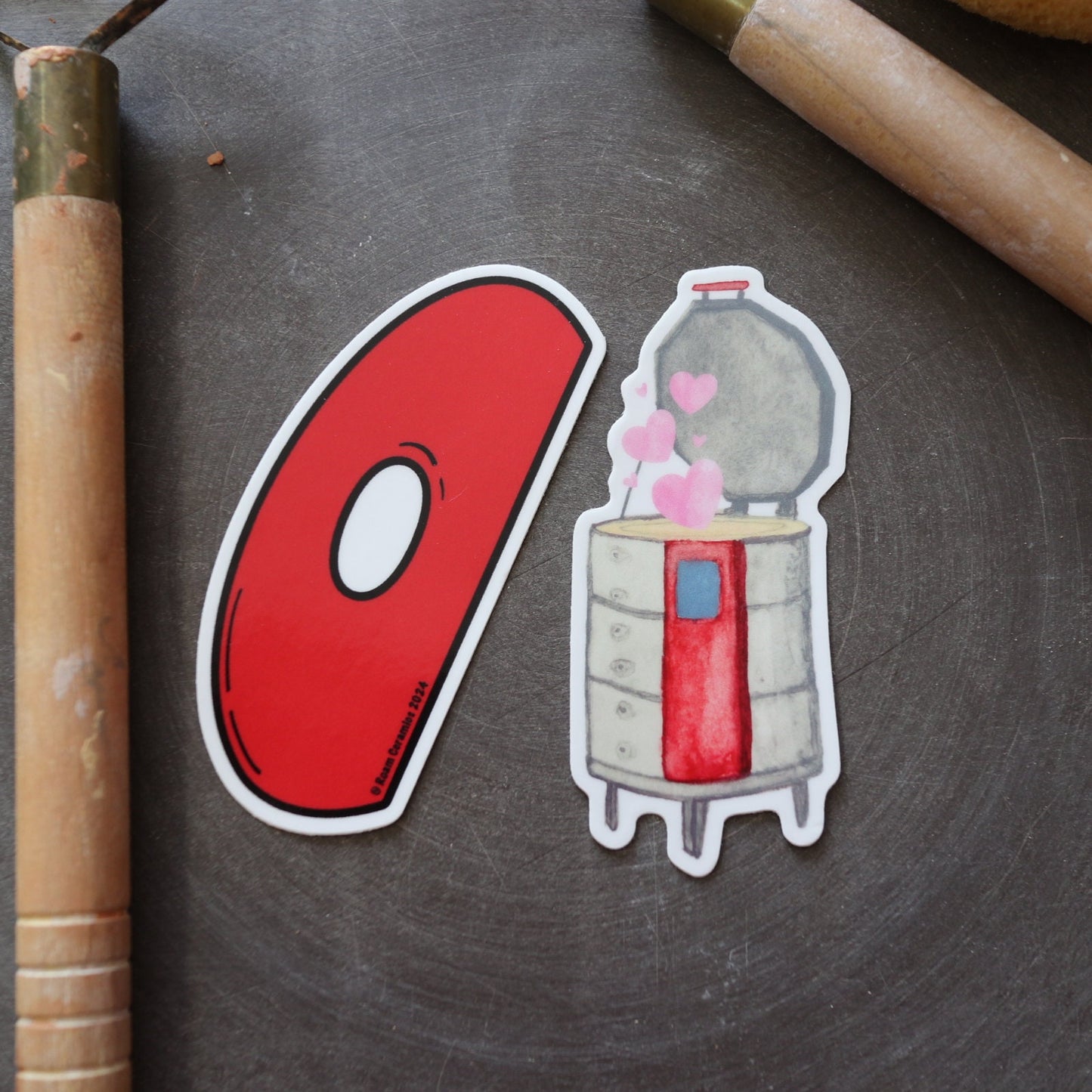 A red, teardrop-shaped vinyl sticker with a white border and a small white circle in the center, placed on a textured grey surface.