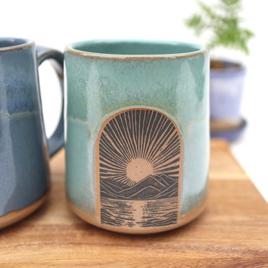 Teal ceramic mug with sun and mountain design on a wooden surface