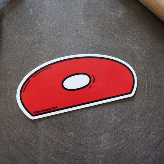 A red, teardrop-shaped vinyl sticker with a white border and a small white circle in the center, placed on a textured grey surface.